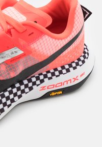 Scarpa da corsa Nike con tomaia di un vivace corallo e un motivo a griglia, dettagli neri e una suola bianca testurizzata con un design a scacchi.