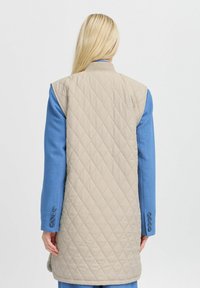 Beige gesteppte Weste im ärmellosen Design, mit einer glatten Textur und einem Diamantmuster. Über einem blauen Hemd mit Knopfdetails getragen.