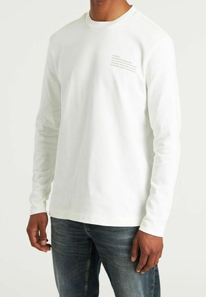 Homme portant une chemise blanche à manches longues avec un petit texte sur le côté supérieur gauche de la poitrine et un jean bleu délavé, debout devant un fond uni.