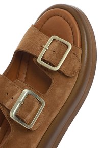 Brown suede sandal featuring two adjustable straps with rectangular metal buckles, rounded toe, and a cushioned brown sole.