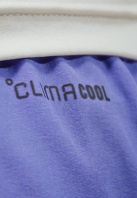 Una tela morada con una textura suave presenta en negro el texto impreso "CLIMA COOL" de manera destacada, situado contra un ribete blanco en la parte superior.