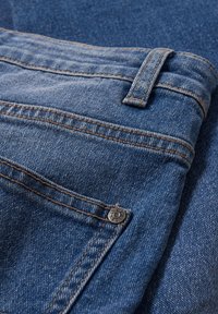 Gros plan d'un jean en denim bleu montrant une poche, un passant de ceinture, une couture orange et un détail de rivet métallique.