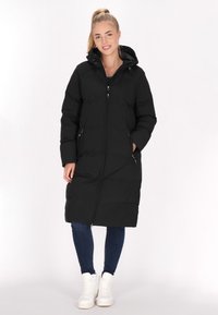 Giacca a vento nera con cappuccio, chiusura con zip e tasche laterali. La giacca ha un design di lunghezza media ed è abbinata a jeans blu e scarpe da ginnastica bianche.