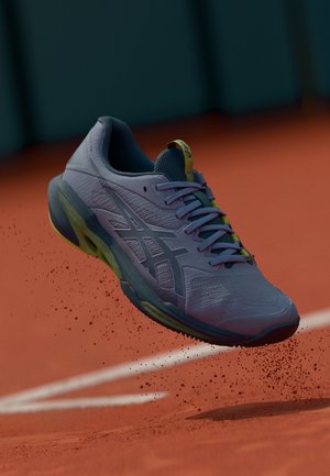 Lichtpaars sportief schoen met een gaasstructuur, gele en teal accenten, en een flexibele zool, verhoogd boven een rode speelvloer.