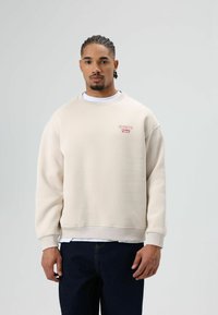 Crèmekleurige trui met een losse pasvorm, geribbelde boorden en zoom, met een klein rood geborduurd logo op de borst, gedragen over een wit shirt.