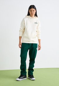 Sudadera crema con cordón ajustable, pantalones de chándal verdes, zapatillas negras de caña alta, con sutiles acentos de logo y un corte relajado.