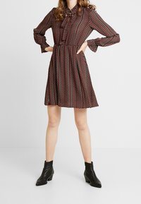 Röd och svart mönstrad klänning med långa ärmar, rosett vid halsen, samlad midja och utsvängd kjol. Bärs med svarta ankelstövlar.