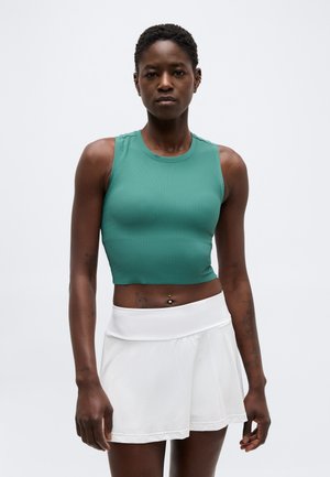 Femme portant un crop top vert sans manches et une jupe blanche, debout devant un fond clair uni.