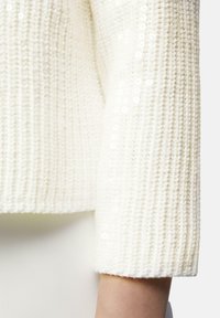 Weißer Strickpullover mit strukturiertem Muster, verziert mit Pailletten entlang des Ärmelabschlusses, mit einem gerippten Bündchen abgeschlossen.