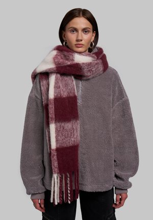 Joven mujer con una gran bufanda de cuadros en color granate y blanco y un suéter gris con cremallera y textura suave, sobre un fondo gris.