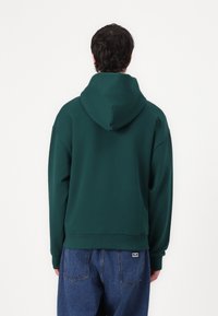 Grønn hettegenser laget av myk stoff, med snorhette, ribbede mansjetter og midje. Bært sammen med blå denimbukser.