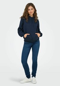 Sweat à capuche bleu marine en coton, avec une poche kangourou à l'avant, une capuche avec cordon et des poignets côtelés. Associé à un jean ajusté et des baskets blanches.