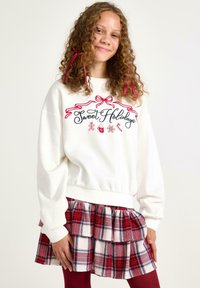 Hvid sweatshirt med "Sweet Holidays" broderi i rød, parret med en lagdelt rød og ternet nederdel. Har krøllet hår og røde bånd detaljer.