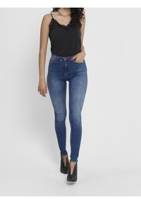 Kvinna som bär en svart topp med spetsdetaljer, högmidjade blå skinny jeans, svarta texturerade klackskor och lager av guldhalsband.