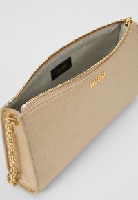 Złota skórzana kopertówka o teksturowanej powierzchni, z zamkiem błyskawicznym, akcentem łańcuszka i zielonym podszyciem. Wewnątrz znajduje się czarna metka z logo.