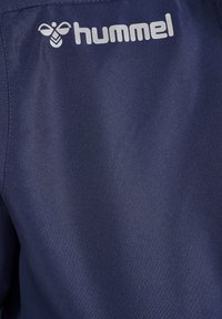 Dunkelblauer Stoff mit glatter Textur, der ein silbernes Logo mit der Aufschrift "hummel" auf der oberen linken Seite zeigt. Einfaches Design, Nahaufnahme.