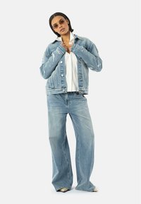 Veste en denim bleu clair avec des détails déchirés, associée à un jean large bleu clair et une chemise blanche en dessous, accessorisé avec des lunettes de soleil.