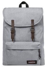 Zaino di tessuto grigio con patta superiore, cinghie in pelle marrone, fibbie metalliche regolabili, tasca frontale e un'etichetta del logo in basso.