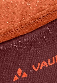 Waterafstotende stof in bruin en oranje kleuren, met een gestructureerd oppervlak en zichtbare waterdruppels. Merklogo gedrukt in oranje.