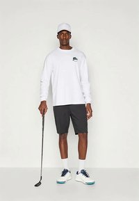 Cămașă albă cu mânecă lungă și logo verde, asociată cu pantaloni scurți negri. Poartă o șapcă albă și pantofi albi cu accente albastre, ținând un club de golf.