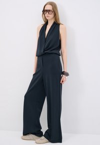 Marinblå jumpsuit med djup V-ringning, avslappnad passform och vida ben. Kombinerad med ljusbeiga sneakers och stora svarta glasögon. Slät textur.