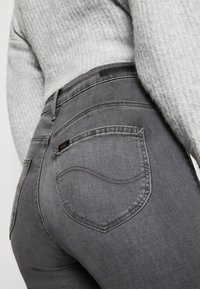 Vaqueros de mezclilla gris con un diseño ajustado, que presentan bolsillos traseros curvados y una etiqueta en el bolsillo derecho. Suéter de punto gris texturizado parcialmente visible.