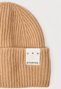 Beige värvitud müts soonilise tekstuuriga, mansetil valge silt musta tekstiga "CPH STUDIOS".