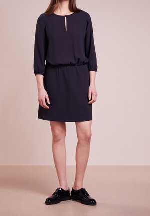 Vrouw met een zwarte jurk tot op de knie, driekwart mouwen en een halslijn met een sleutelgat, gecombineerd met zwarte lakleren veterschoenen.