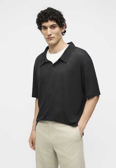 Νέος άνδρας με σγουρά σκούρα μαλλιά που φορά χαλαρό μαύρο κοντομάνικο πουκάμισο πάνω από λευκό φανελάκι και ανοιχτό μπεζ παντελόνι, στέκεται με το ένα χέρι στην τσέπη.