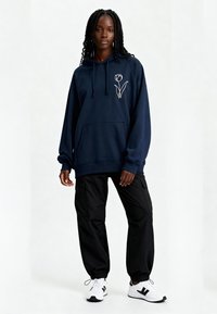 Sweat à capuche bleu marine avec une broderie florale blanche, poche kangourou, associé à un pantalon cargo noir et des baskets blanches. Coupe décontractée et capuche avec cordon de serrage.