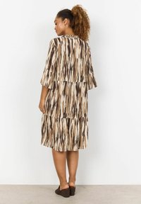 Abito fluente e a più strati in beige e marrone con un motivo a zigzag; presenta maniche corte e un collo rotondo. Indossato con sandali con tacco a cuneo marroni.