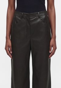 Personne portant un pantalon en cuir noir taille haute à jambes larges avec poches avant et passants de ceinture, associé à un haut noir sans manches.