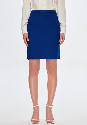 Femme portant une jupe crayon bleu roi longueur genou, un chemisier crème à manches longues et des talons blancs à bout ouvert avec bride à la cheville, debout devant un fond blanc.