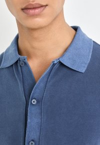 Navy polo shirt with a contrasting light blue collar, featuring a button-up neckline and textured fabric. Close-up view focuses on details.
