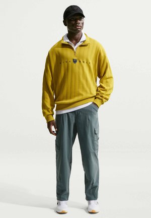 Man met zwarte pet, gele Nike Golf trui, witte polo, grijze cargobroek en witte sneakers, staand met één hand in de zak.