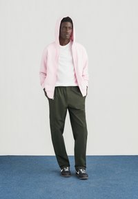 Jasnoróżowa bluza na suwak na białej koszulce, zestawiona z ciemnozielonymi spodniami typu cargo. Model stoi na niebieskim dywanie na tle szarej ściany.