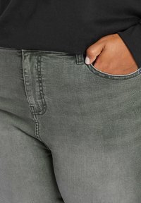 Jeans de mezclilla verde oscuro con costuras visibles, bolsillo delantero y una textura suave. Una mano descansa en el bolsillo lateral.