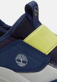 Baskets bleu marine et gris clair avec une sangle jaune, en tissu, col rembourré, et un logo arbre sur le côté.