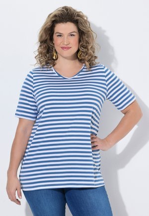 Kobieta z kręconymi włosami w niebiesko-białej pasiastce koszulce z krótkim rękawem i niebieskich dżinsach, stojąca z jedną ręką na biodrze, uśmiechająca się na białym tle.