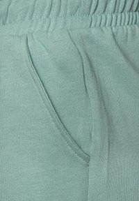 Gaiši teal audums ar mīkstu tekstūru, piedāvājot elastīgu jostasvietu un sānu kabatu, ar vienkāršu adījuma rakstu un redzamām šuvēm.