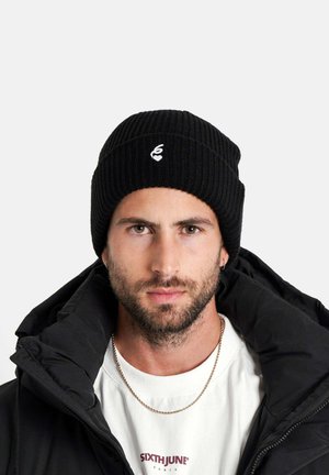 Bonnet en tricot côtelé noir avec un petit logo blanc. Le modèle porte une doudoune noire et une chemise blanche avec du texte en-dessous.