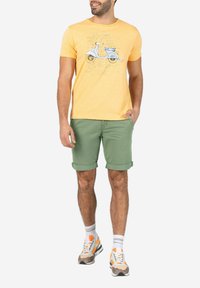 Gelbes Baumwoll-T-Shirt mit einem Scooter-Graphic; kombiniert mit hochgekrempelten grünen Shorts und mehrfarbigen Turnschuhen, die eine strukturierte Sohle haben.