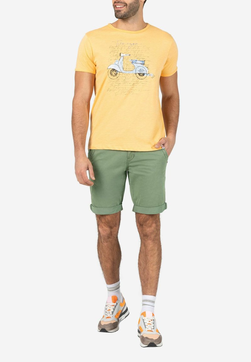 Gelbes Baumwoll-T-Shirt mit einem Scooter-Graphic; kombiniert mit hochgekrempelten grünen Shorts und mehrfarbigen Turnschuhen, die eine strukturierte Sohle haben.