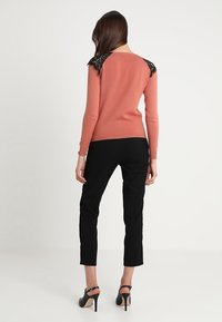 Světle hnědý svetr s černými krajkovými detaily na ramenech, kombinovaný s elegantními černými kalhotami a vysokými podpatky, vyfotografováno zezadu.