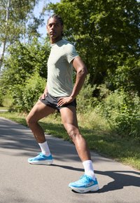 Athletic man in a light green shirt and black shorts, wearing blue HOKA running shoes and white socks, posing on a path with greenery nearby.