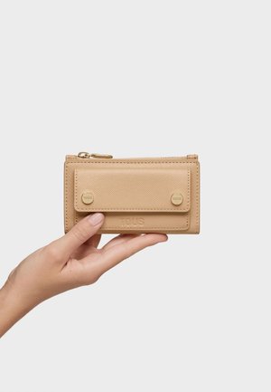 Mano sosteniendo una cartera de cuero beige con solapa frontal asegurada por dos botones dorados y un compartimento con cremallera en la parte superior.