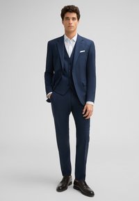 Marineblaues Dreiteiliges Anzug mit einreihigem Jacket, schmal geschnittenen Hosen und einem weißen Hemd mit Kragen; ergänzt durch schwarze Anzugschuhe.