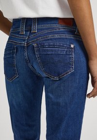 Jeans de mezclilla azul oscuro con un diseño ajustado, que cuentan con dos bolsillos traseros, costuras naranjas y una etiqueta de cuero marrón en la cinturilla.