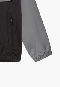 Dwukolorowa kurtka z czarnym korpusem i szarymi rękawami. Cechy to zapinana na zamek kieszeń oraz elastyczne mankiety. Wykonana z lekkiego, wodoodpornego materiału.