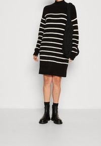 Abito maglione a righe bianche e nere con maniche lunghe e collo alto, abbinato a una borsa a maglia nera e stivaletti neri.
